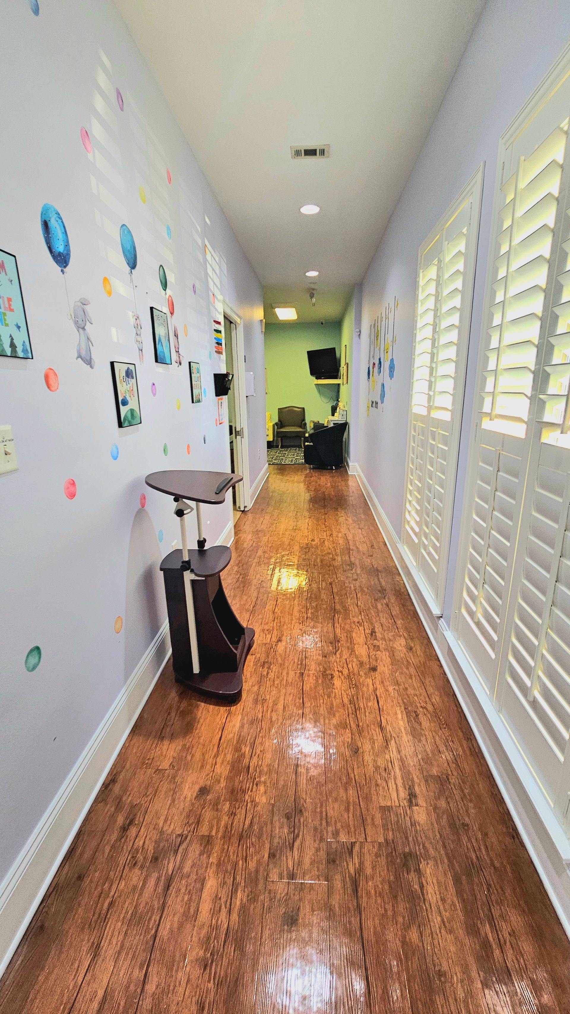 Long hallway with light purple walls, wood floor, and white shutters.  Artwork on the left wall.