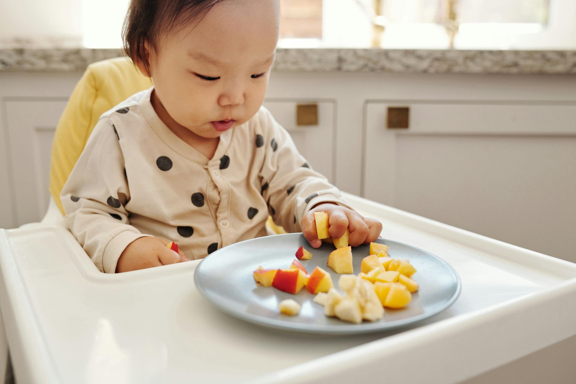 Child in a high chair eating fruit from a plate in a kitchen.