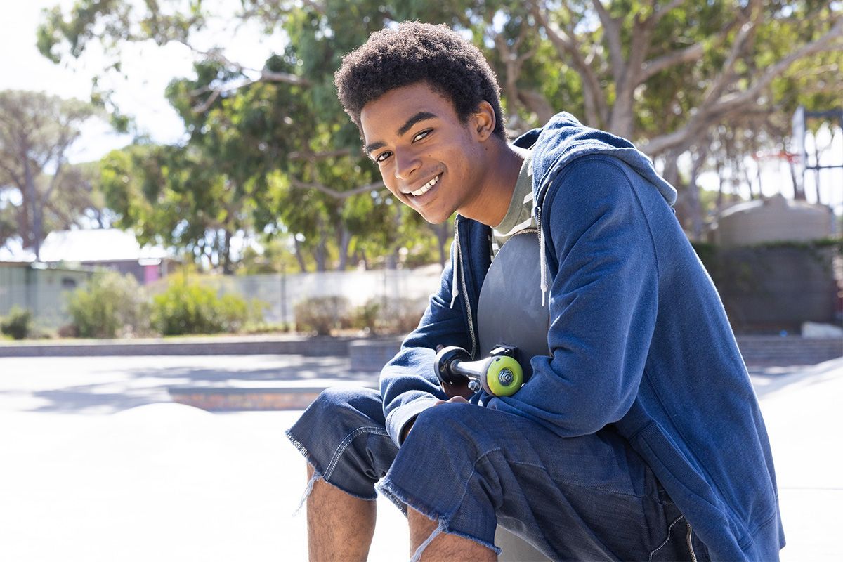 Young person smiles, seated, holding skateboard in park. Wearing blue hoodie, denim shorts.