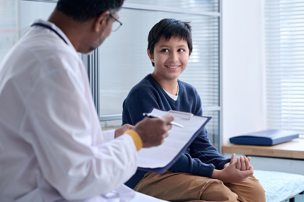 Boy smiles at doctor in exam room; doctor holds clipboard, wearing a white coat.