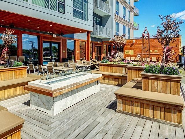 Outdoor patio with fire pit, seating, and wooden planters in front of a modern building at The Walkway Apartments.