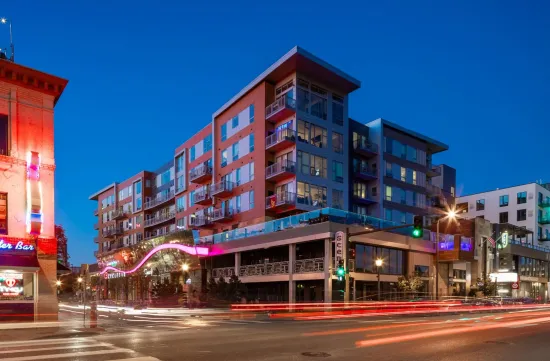 Multi-story apartment building with street-level restaurants at dusk; blurred car lights, city lights, and clear blue sky.
