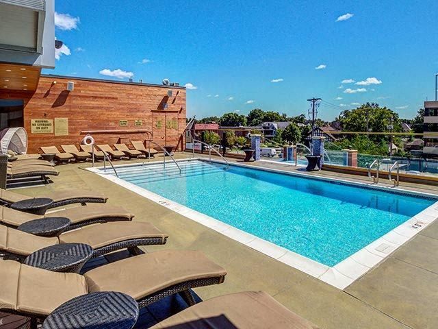 Rooftop pool with lounge chairs, blue water, and brick building under a clear sky The Walkway Apartments.