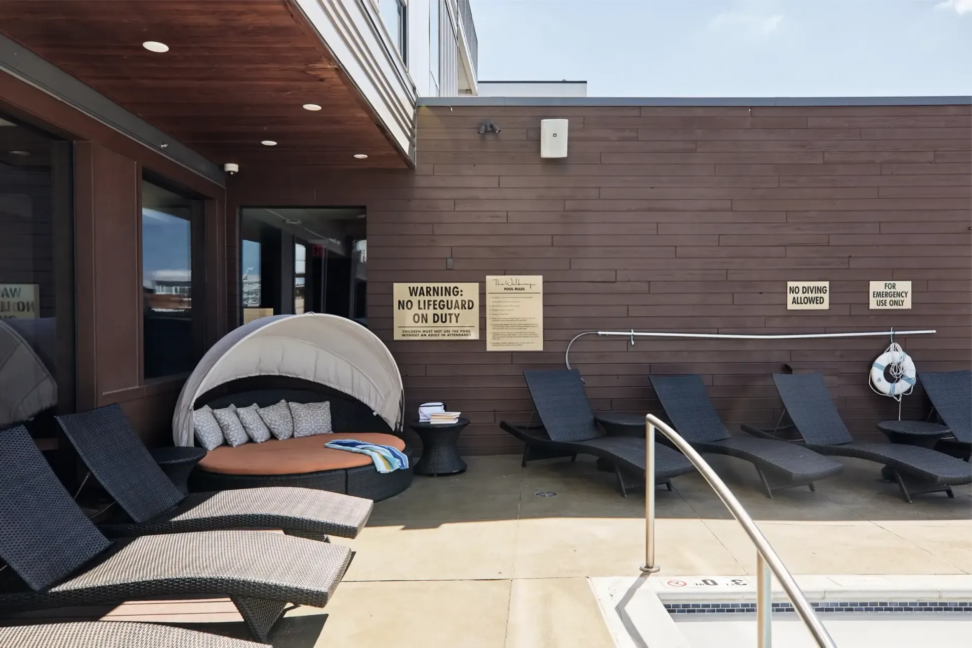 Poolside lounge area with wicker chaise lounges, a curved canopy daybed, and signs on a wood-paneled wall at The Walkway, which offers apartments for rent in Minneapolis, MN.