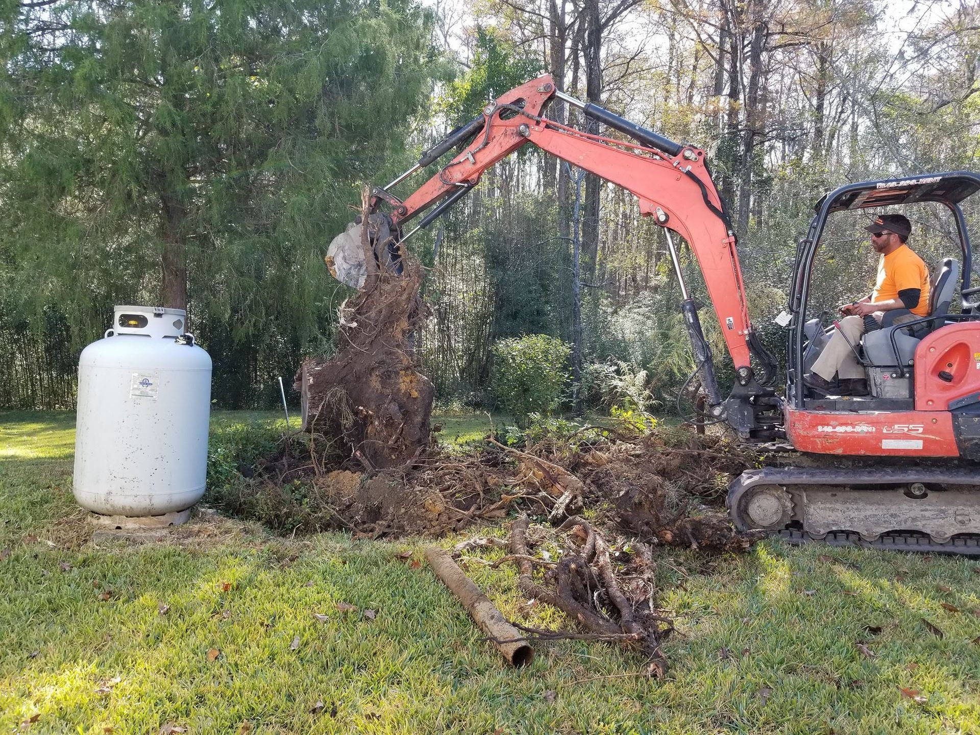 A man is driving an excavator in a yard next to a propane tank.