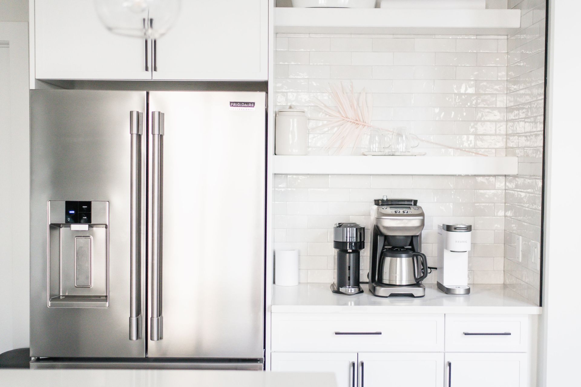 Stainless steel refrigerator next to a coffee station in a white kitchen.