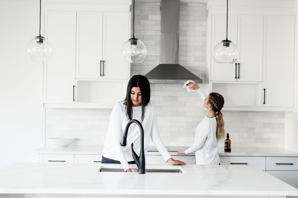 Two women in white kitchen cleaning, one at sink, other near range hood.