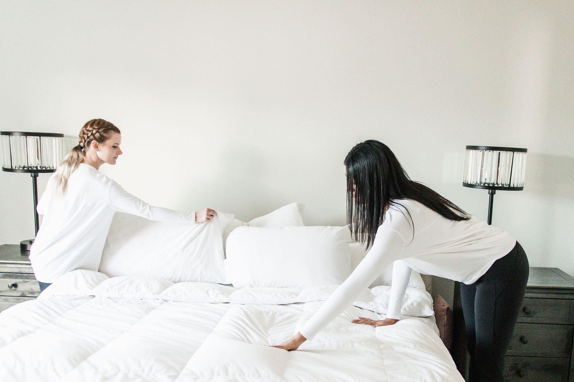 Two women making a white bed in a bedroom.