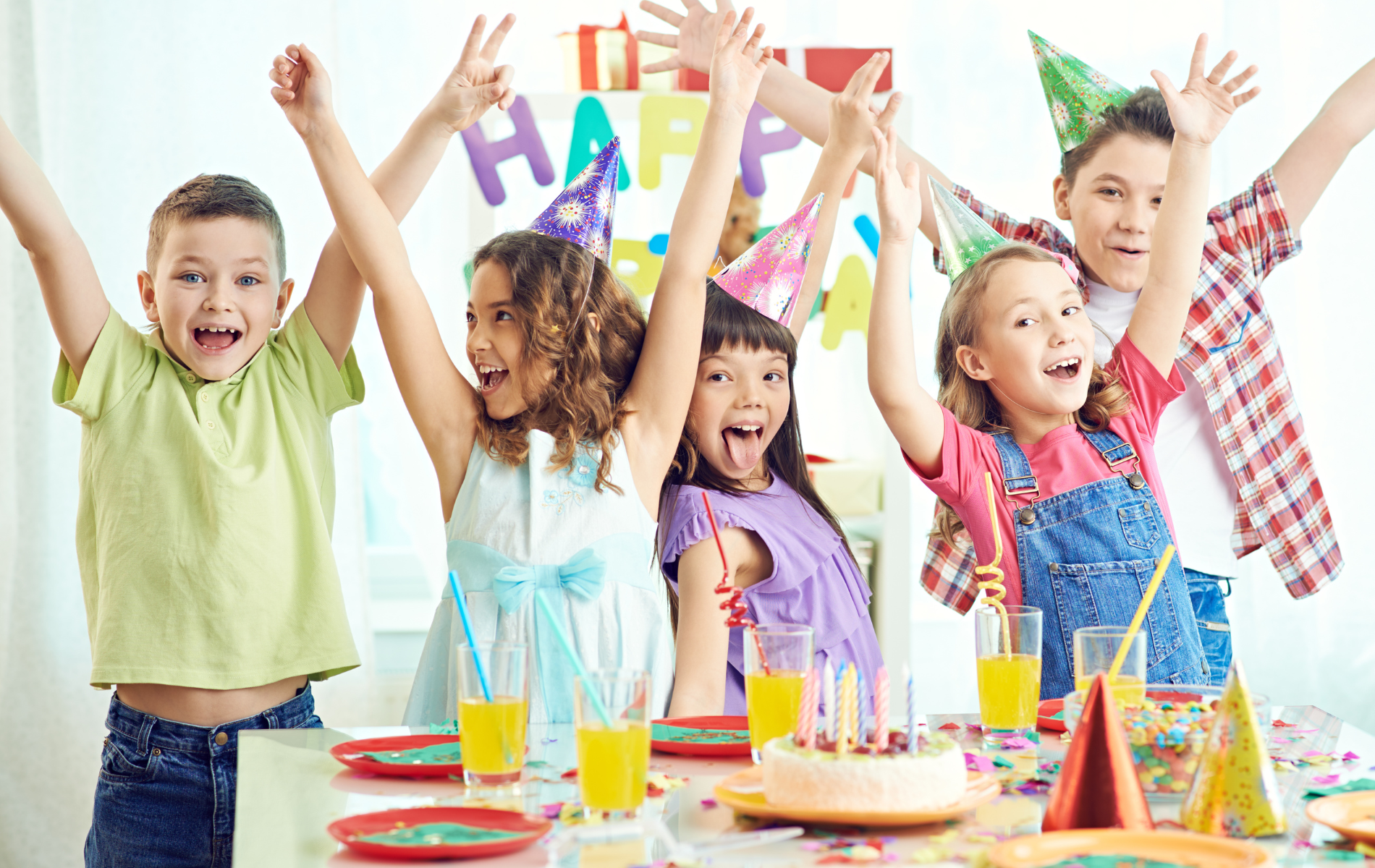 A group of children are sitting at a table with their arms in the air at a birthday party.