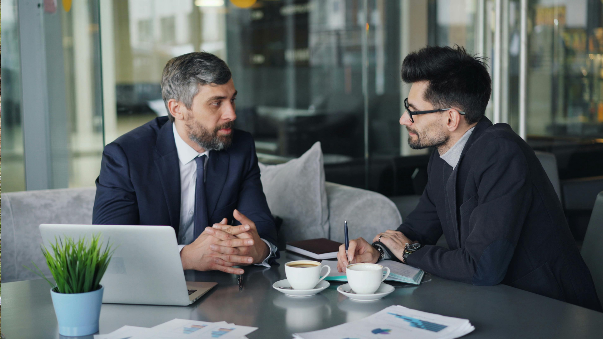 Two men in suits having a discussion at a table with coffee, a laptop, and documents in a cafe.