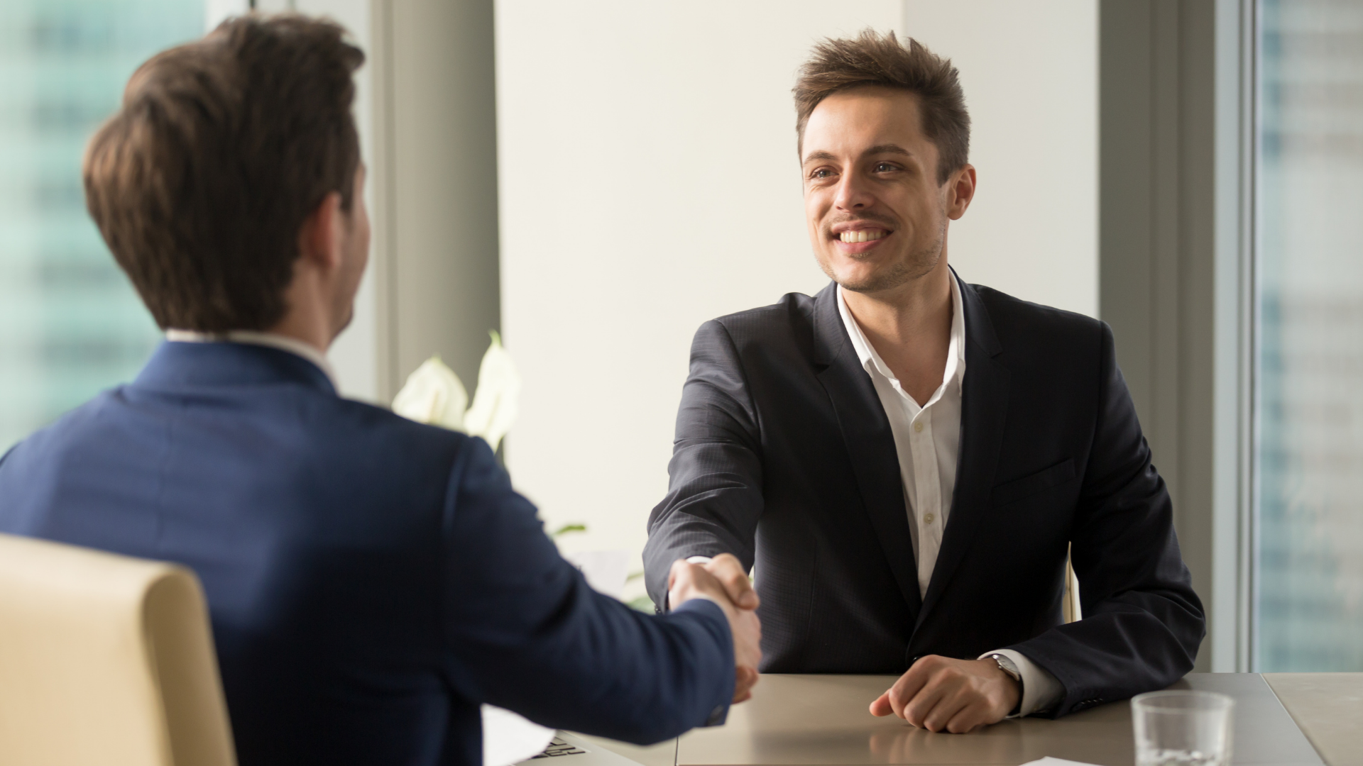 Two men in suits shaking hands at a table, smiling, in an office.