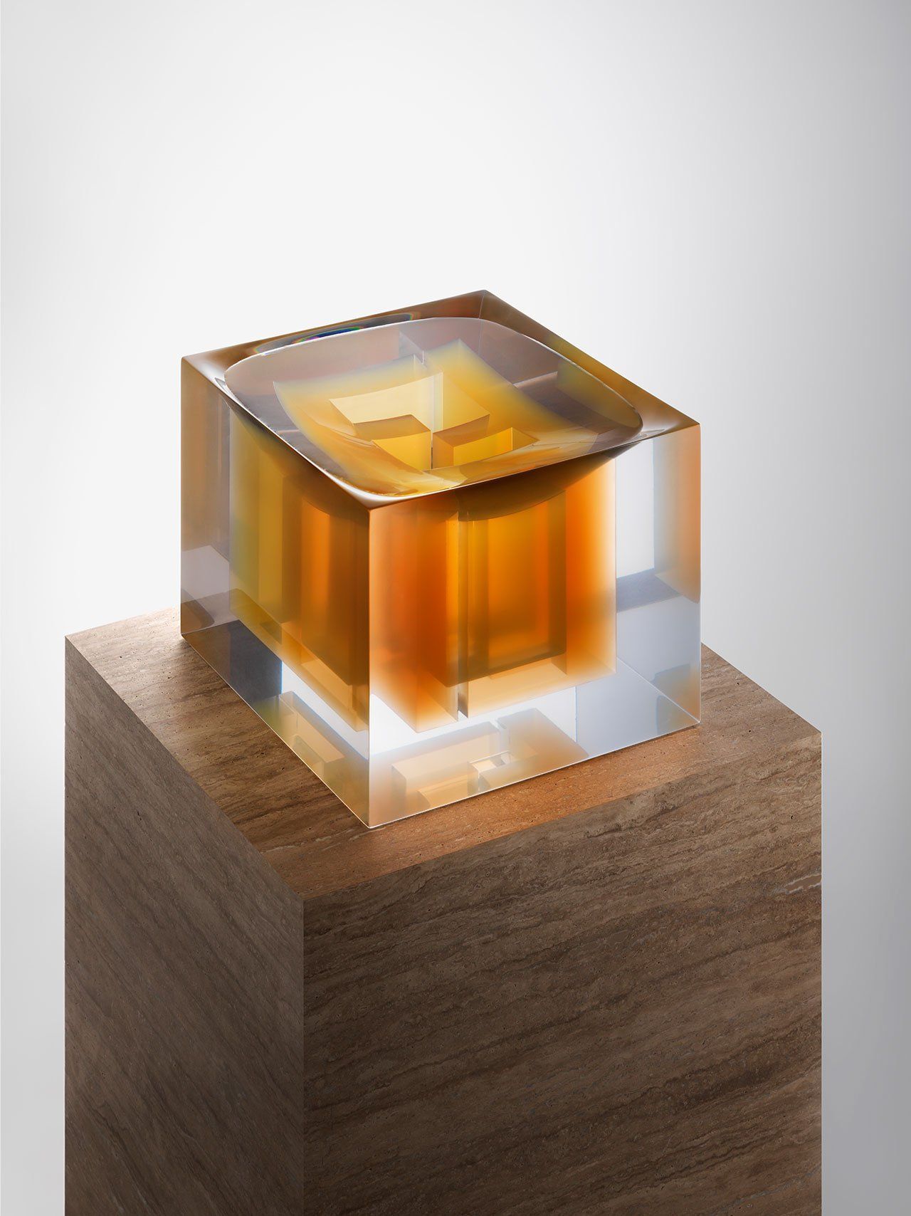 Sculptuur van oranje en helder glas op een houten blok, tegen een witte achtergrond.