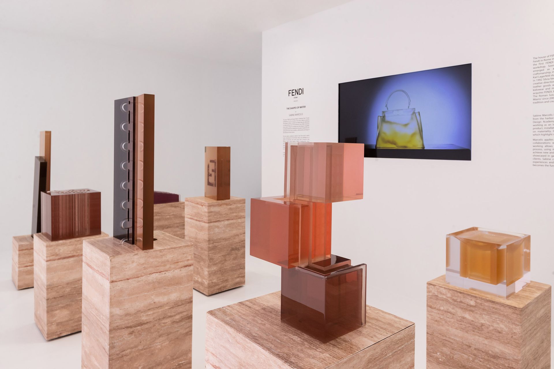 Kunstinstallatie: Geometrische sculpturen in tinten bruin en amber op beige sokkels, met een videoscherm aan de muur.