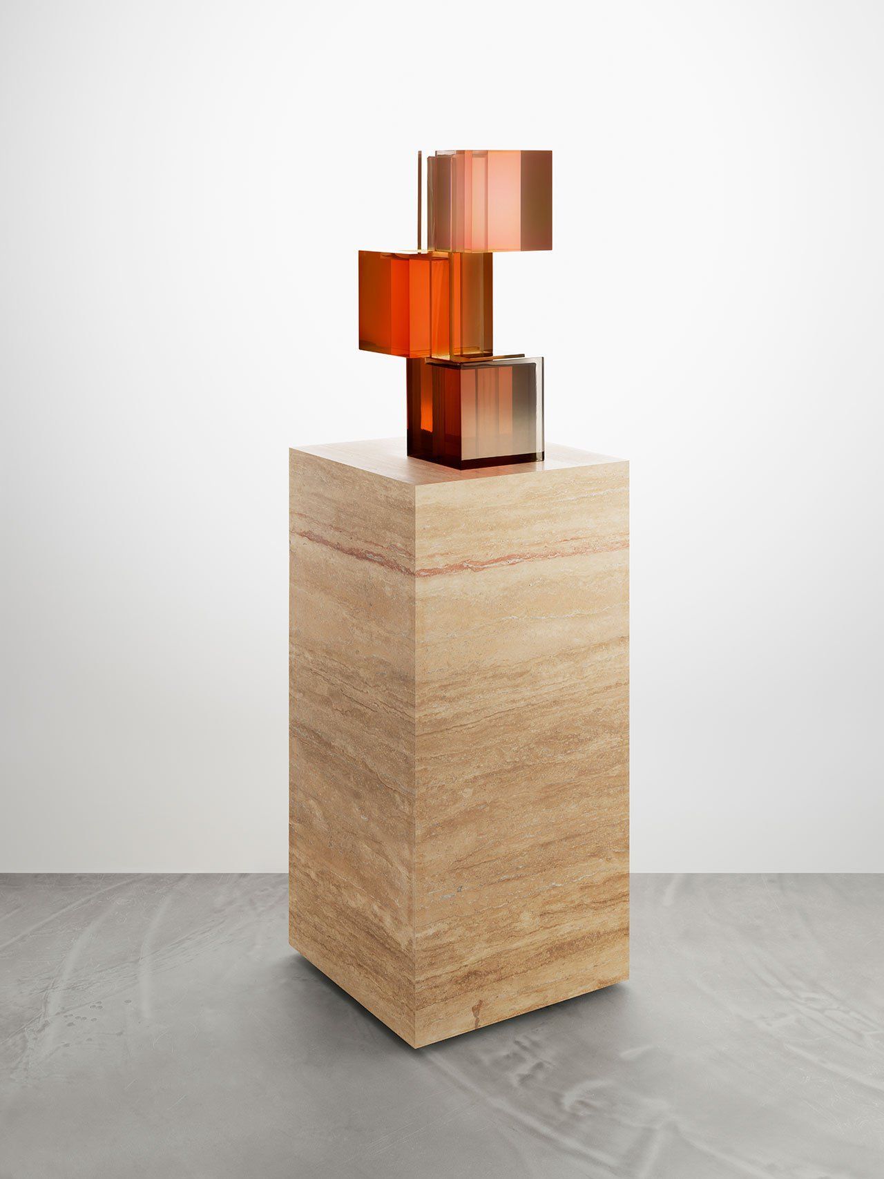 Gestapelde koperen kubussen op een bruine stenen sokkel, tegen een witte muur en grijze vloer.