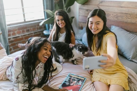 Three people pose for a selfie on a bed with a black and white cat while lounging with a magazine in a cozy bedroom.