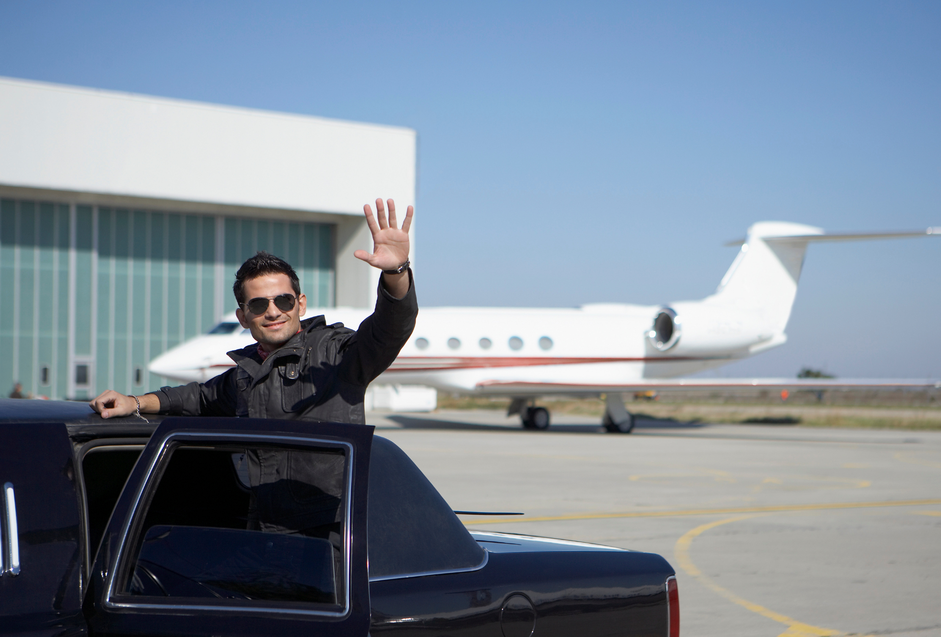 A man is waving from the back of a limousine in front of a private jet.