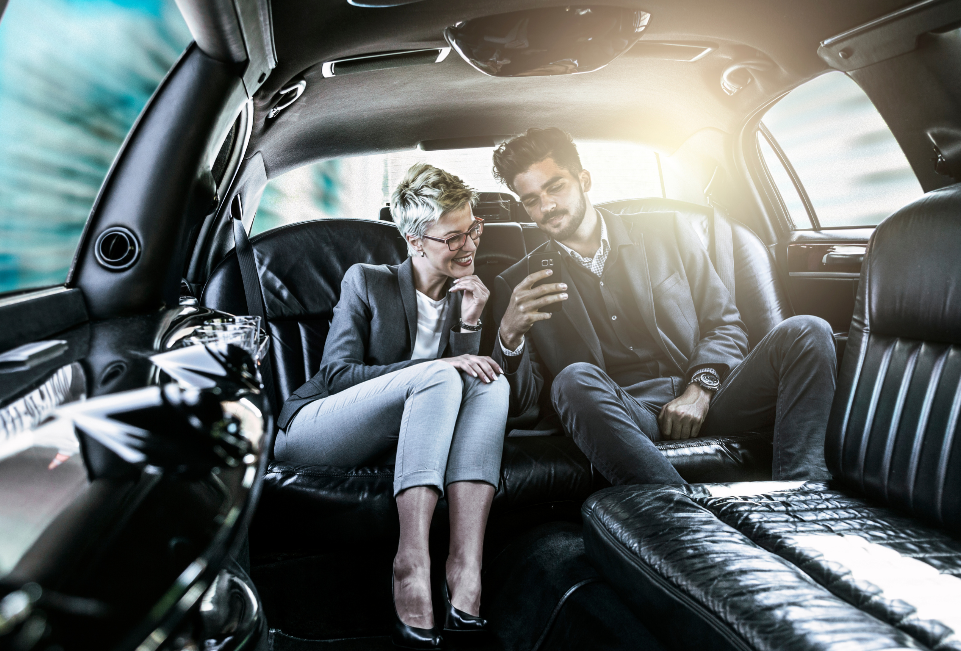 A man and a woman are sitting in the back seat of a limousine looking at a tablet.