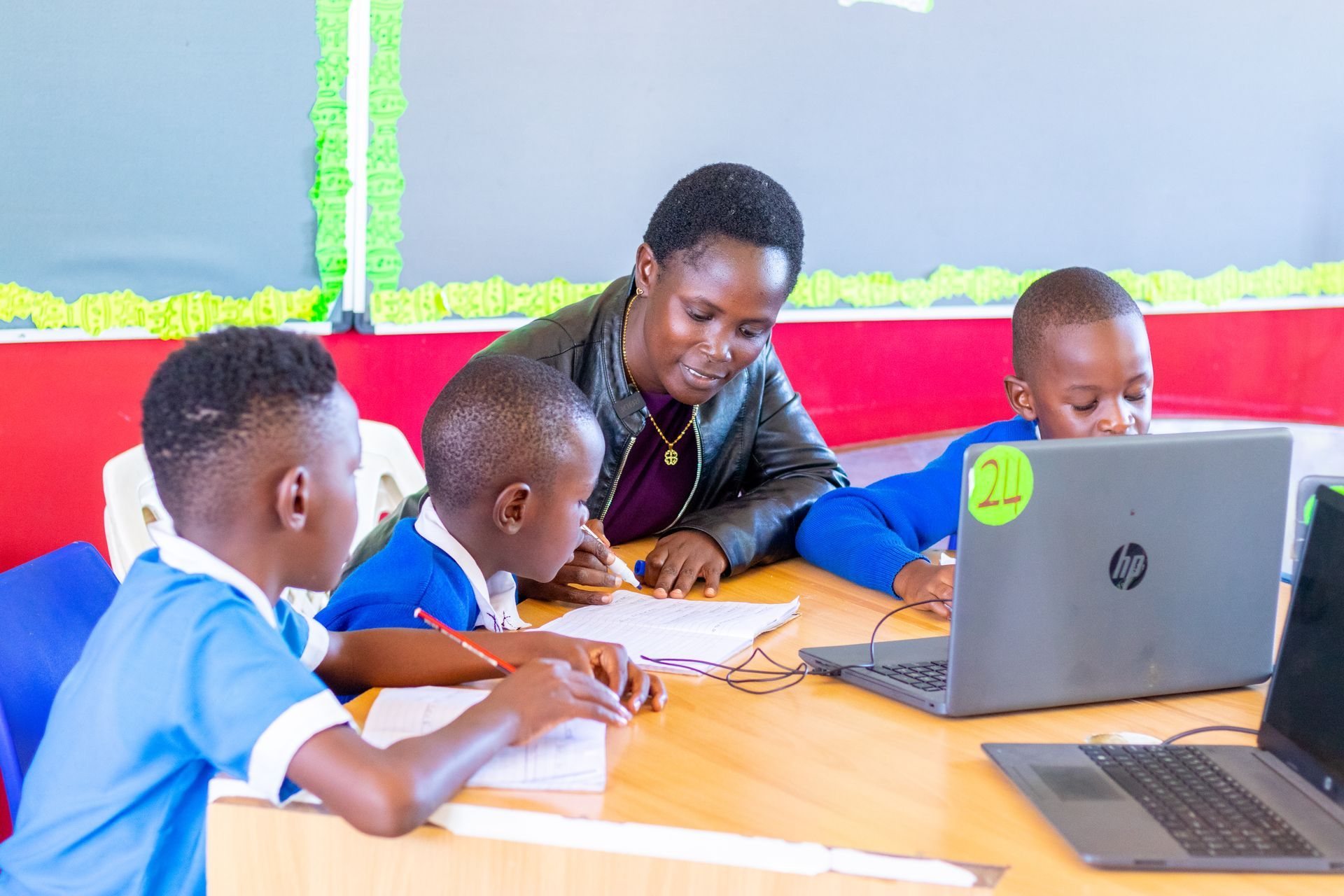 A group of children are sitting at a table with laptops and a teacher.
