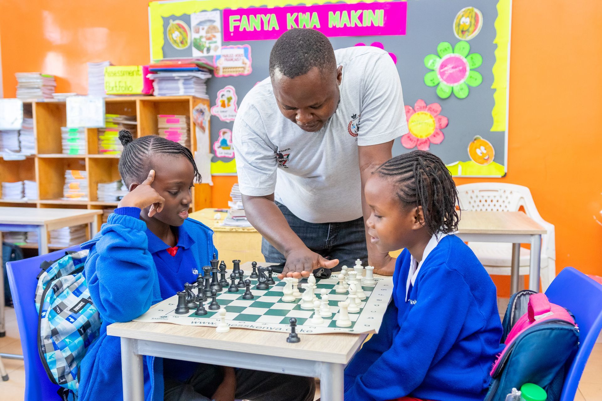 A man is teaching two young girls how to play chess in a classroom.