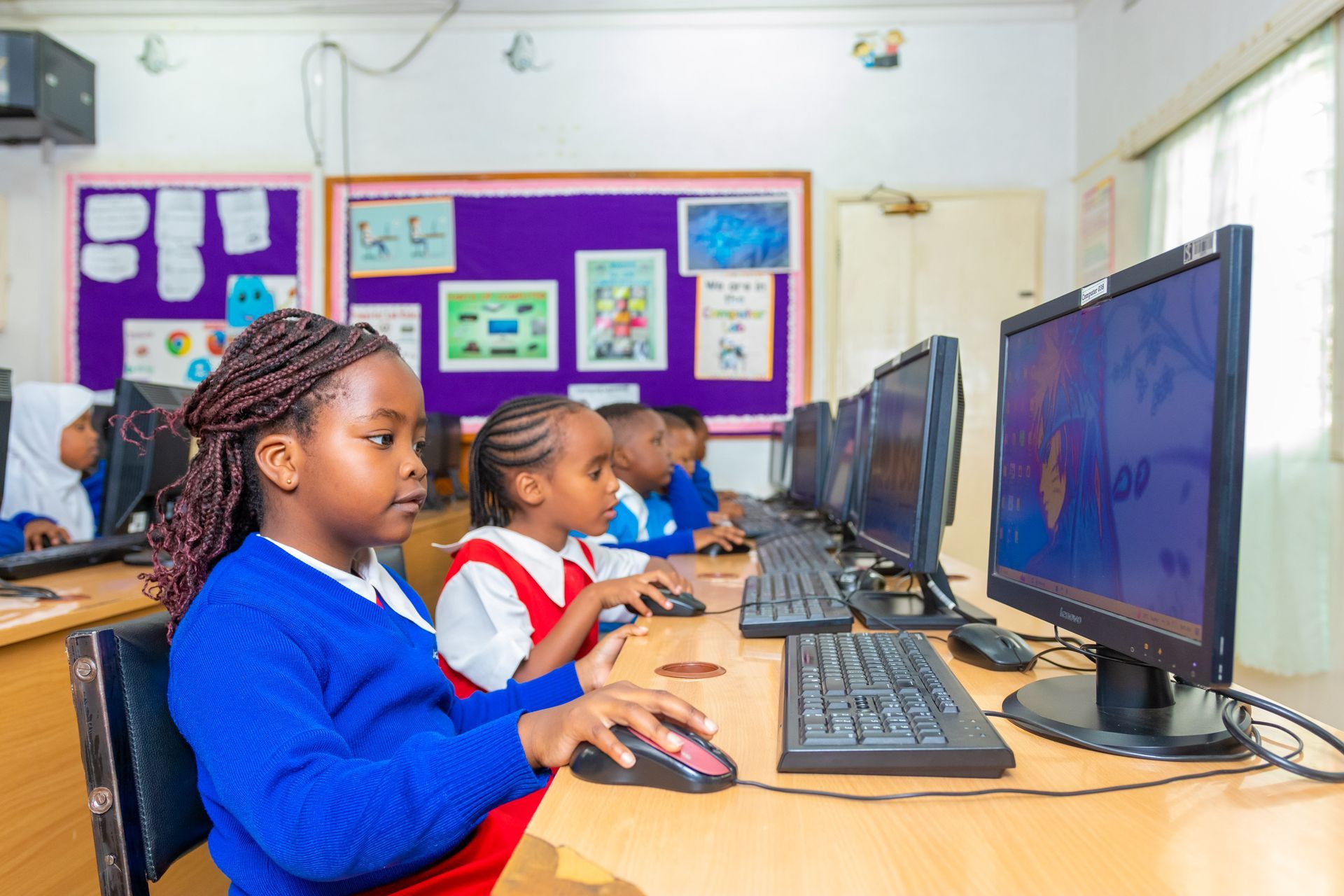 A group of children are sitting in front of computer monitors in a classroom.