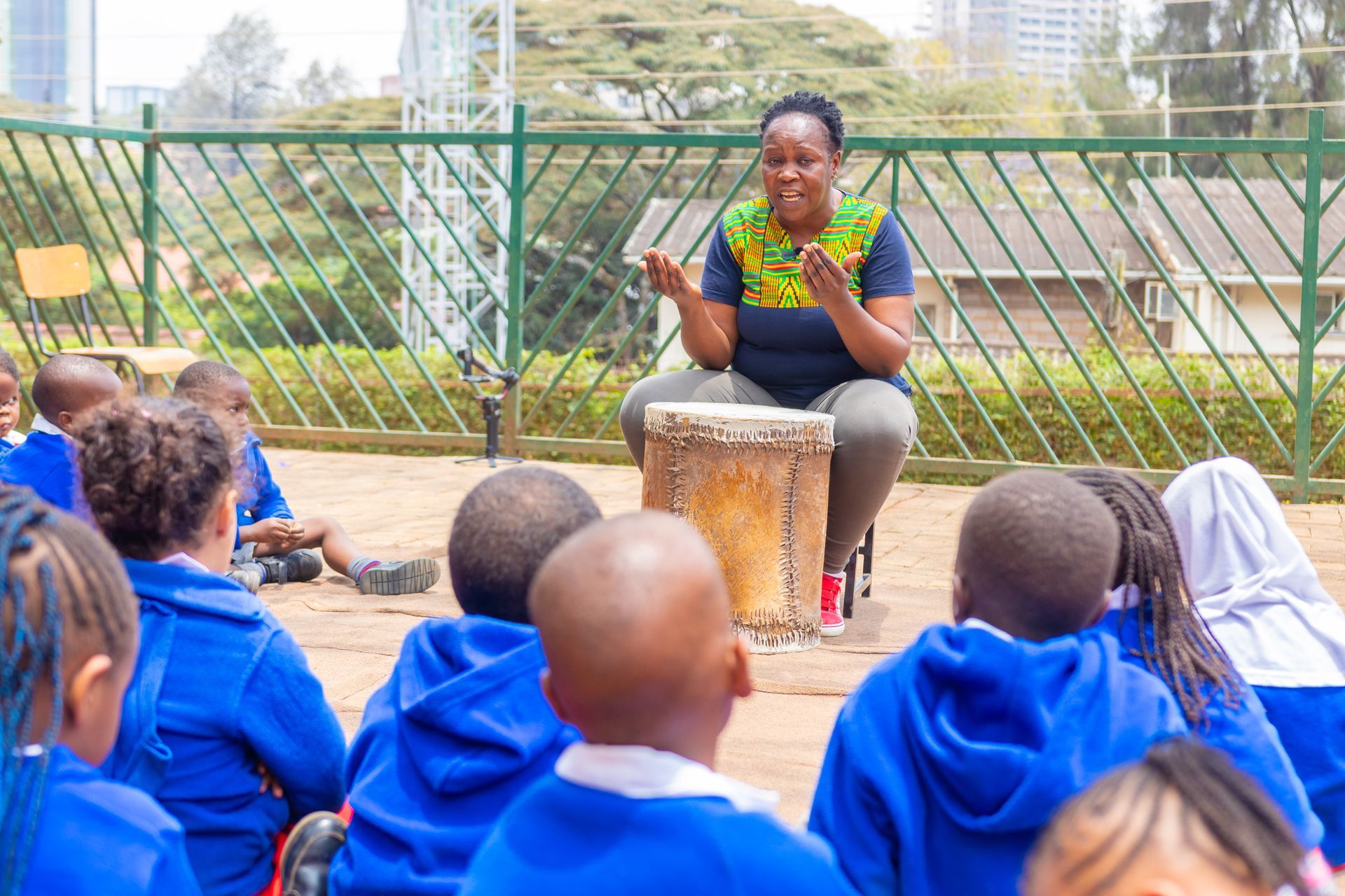 A woman is sitting on a drum talking to a group of children.