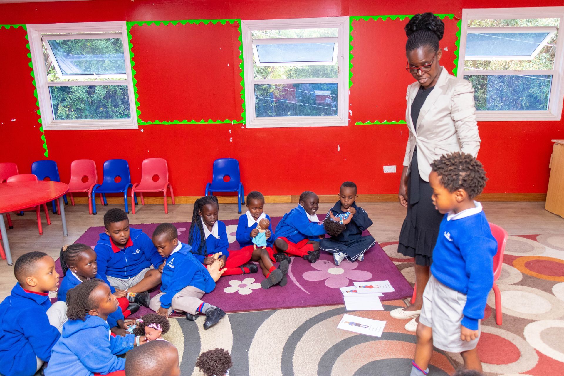 A teacher is standing next to a group of children sitting on the floor in a classroom.