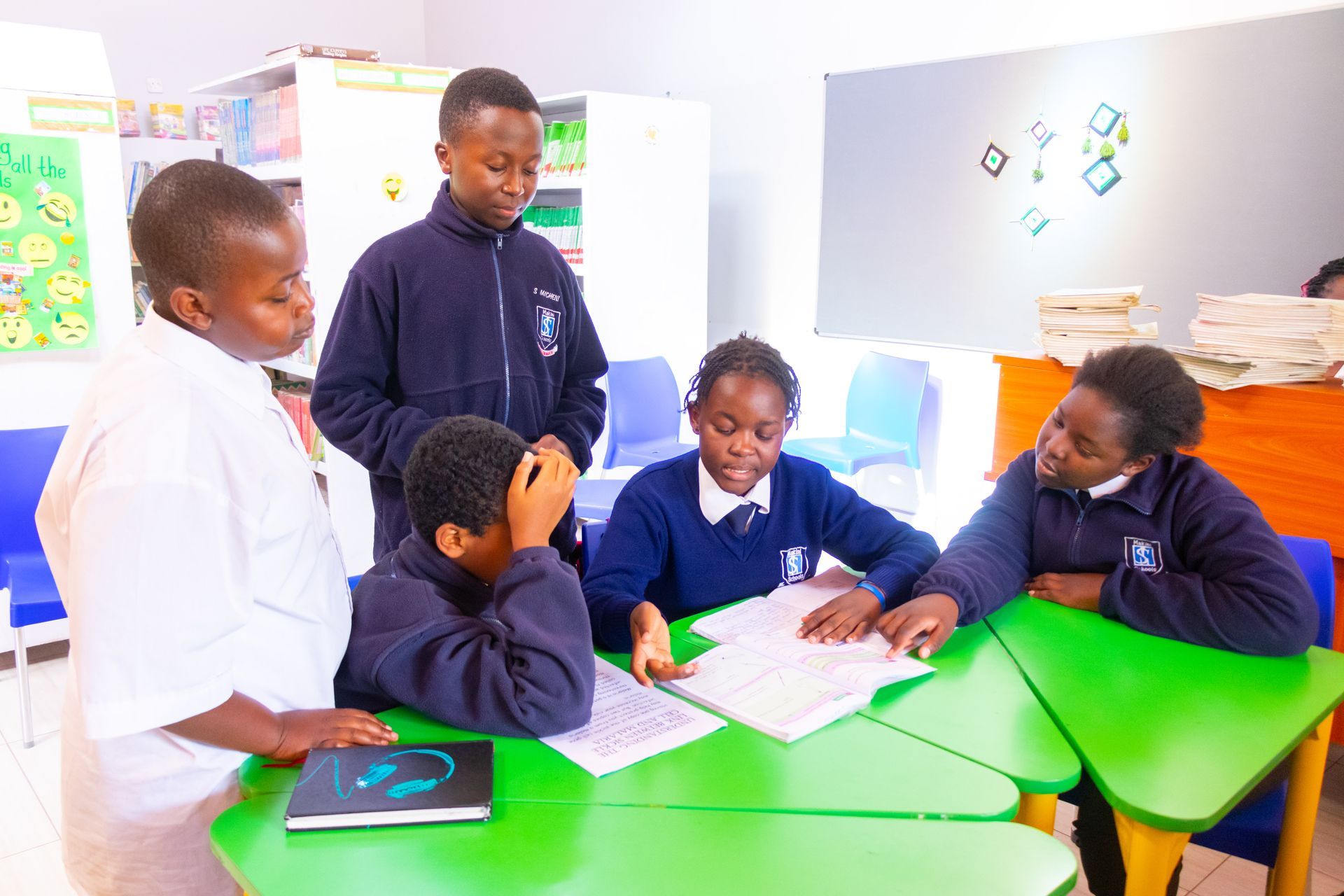 Schoolchildren gather around a green table, looking at papers. Some wear blue uniforms, others white. One boy holds his head.