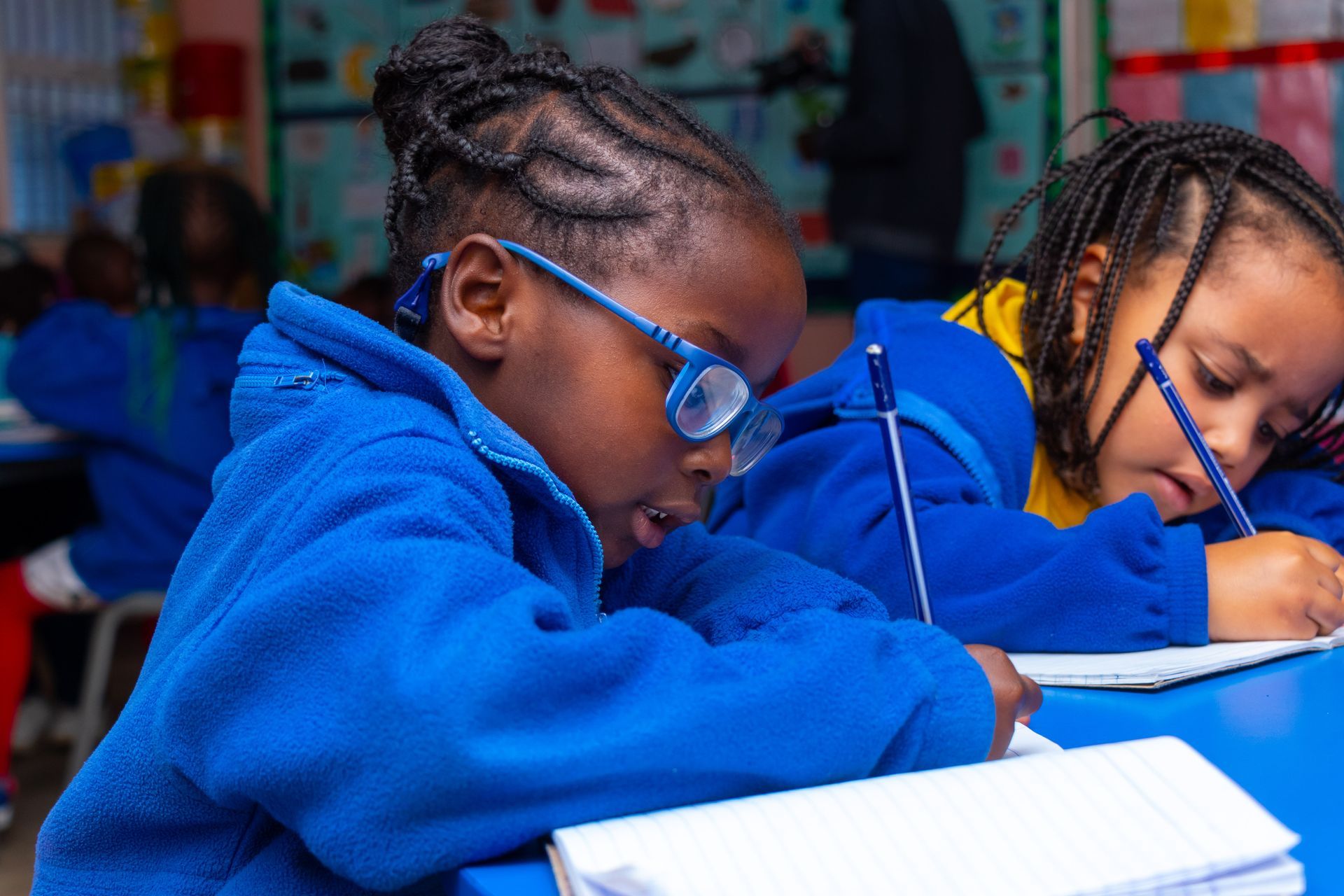Two young girls are sitting at a table in a classroom writing in notebooks.