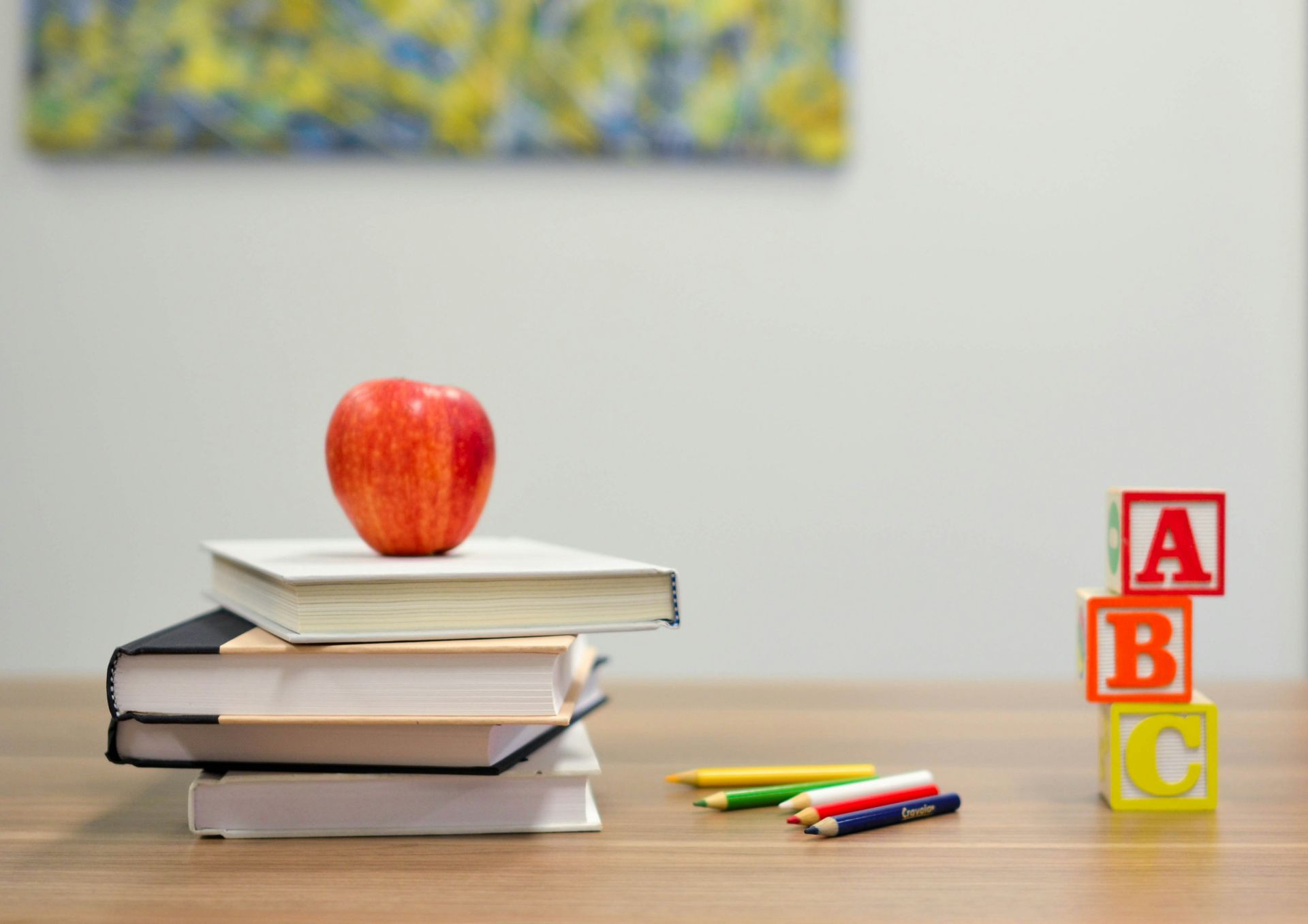 Stack of books topped with apple. ABC blocks and colored pencils on a wooden desk.