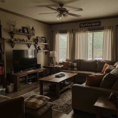 Cozy living room with sectional sofa, wooden furniture, and shelves with decorations.  A TV sits on a wooden console.  Neutral colors, warm lighting.