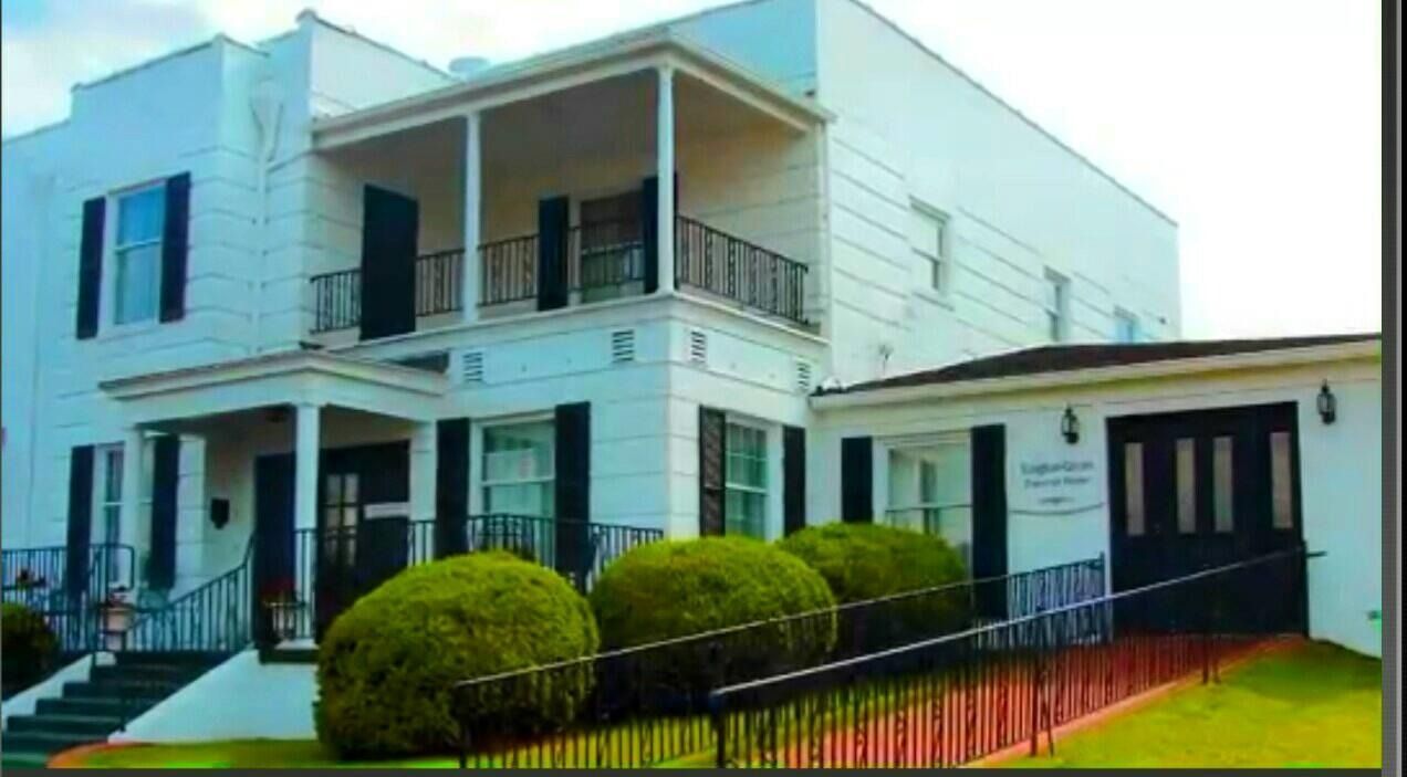 White two-story building with black shutters and railings, entrance ramp, three round green bushes.