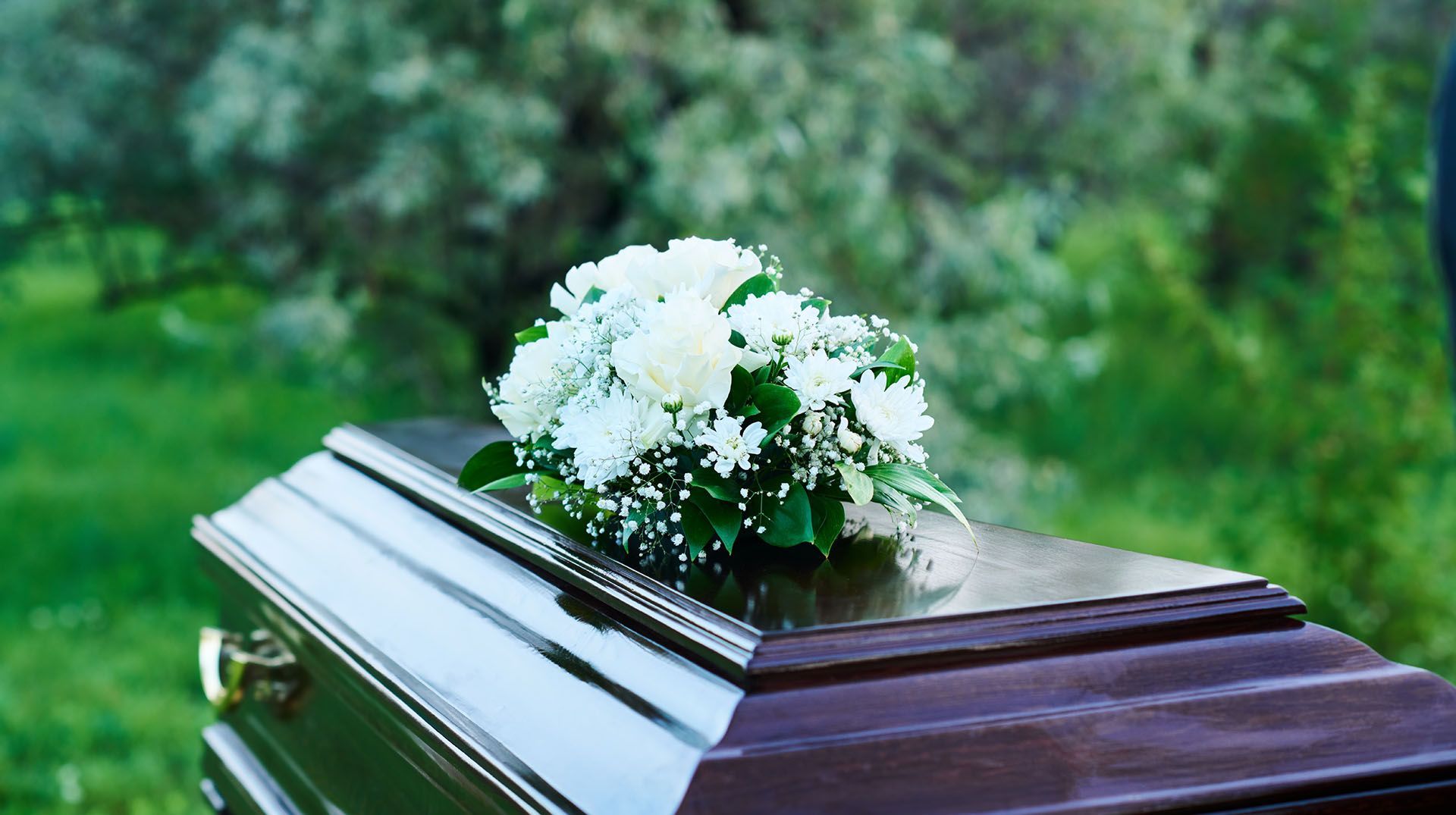 Coffin with white flower arrangement outdoors on green grass.