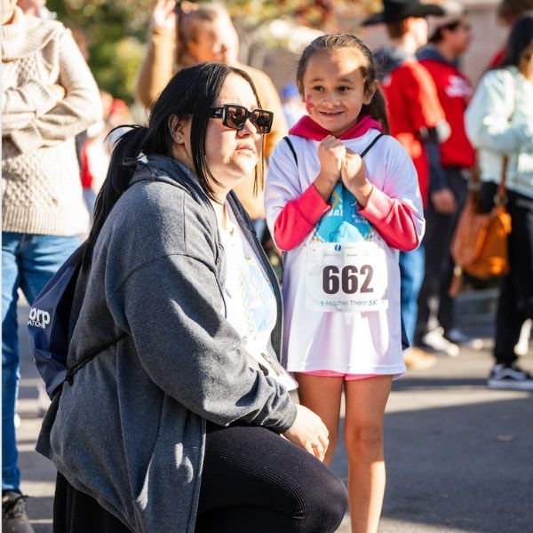 Woman in sunglasses sits next to smiling girl in a race bib; they are outdoors among other people.