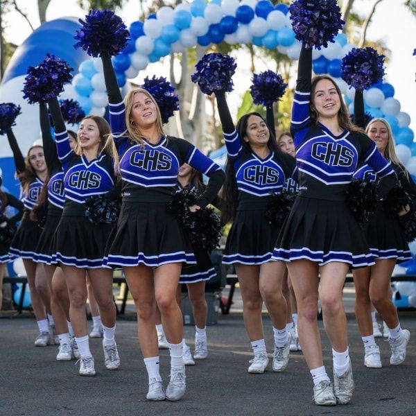 Cheerleaders in black and blue uniforms with pom-poms perform in front of an arch of blue balloons.