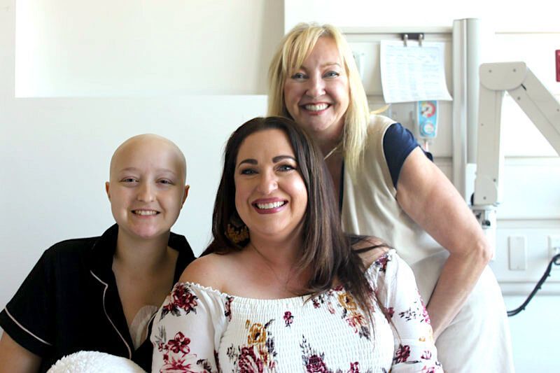 Three smiling women, two with long hair, one bald. Hospital room setting.