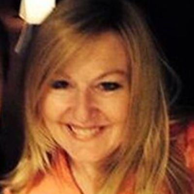 Blond woman with a warm smile, wearing an orange top, indoors.