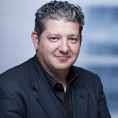 Man with gray curly hair wearing a black shirt and dark patterned jacket, smiling, against a blurred background.