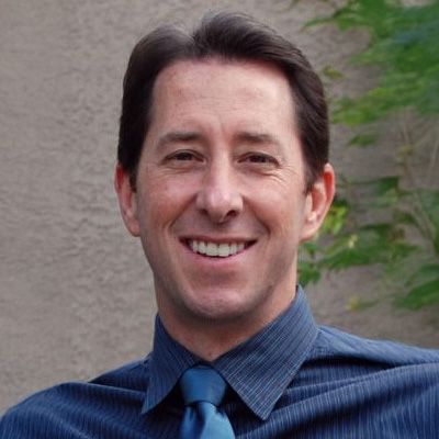 Man with dark hair, smiling, wearing a blue dress shirt and tie, outdoors.