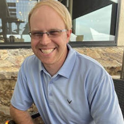 Man with blond hair wearing glasses and a light blue polo shirt smiles outside.