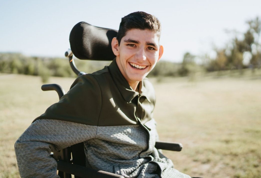 Smiling person in wheelchair outdoors. Green jacket, gray shirt, and sunny background.