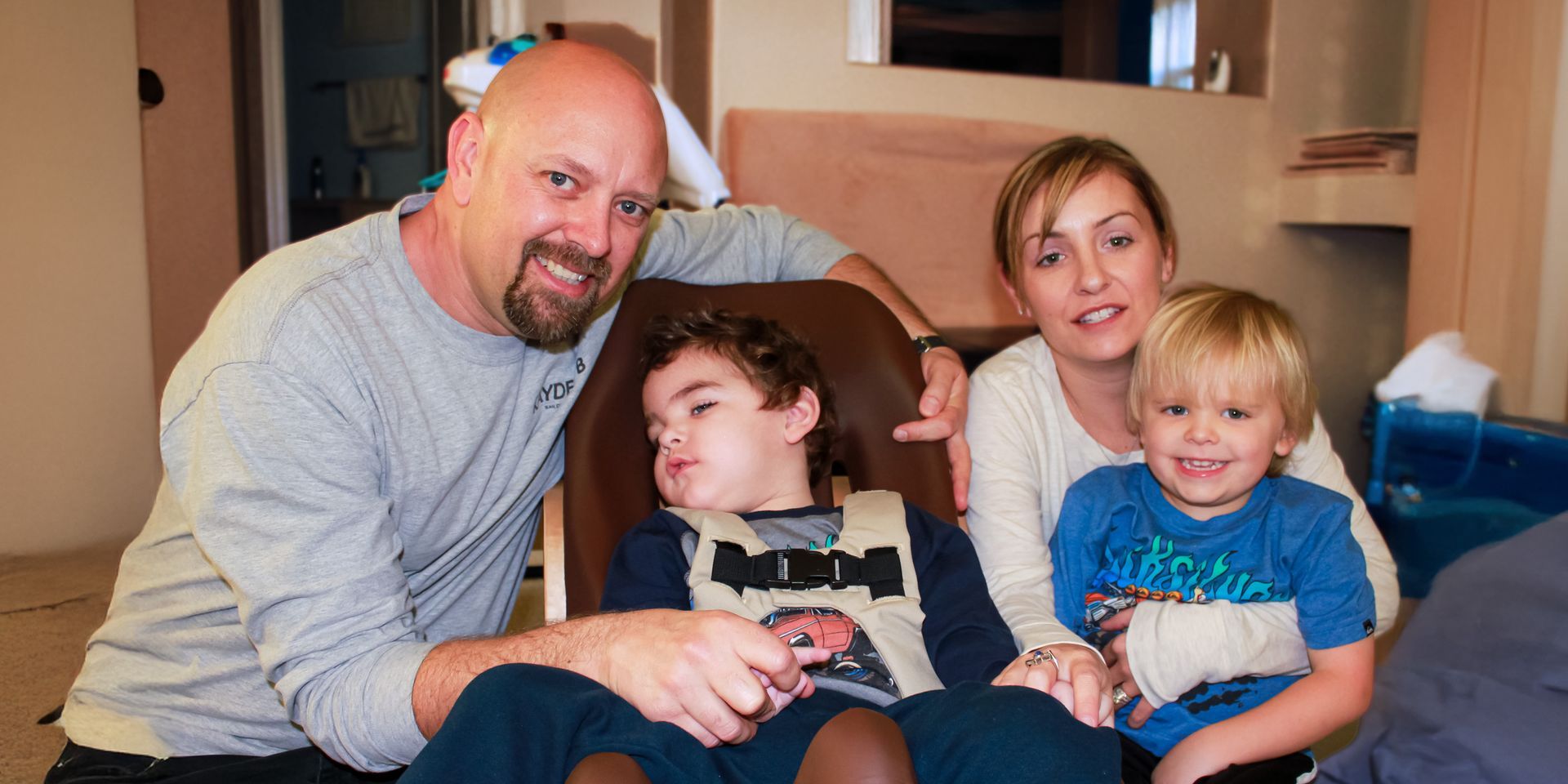Family of four smiling at the camera; a man, woman, and two young boys, one in a wheelchair with a harness.