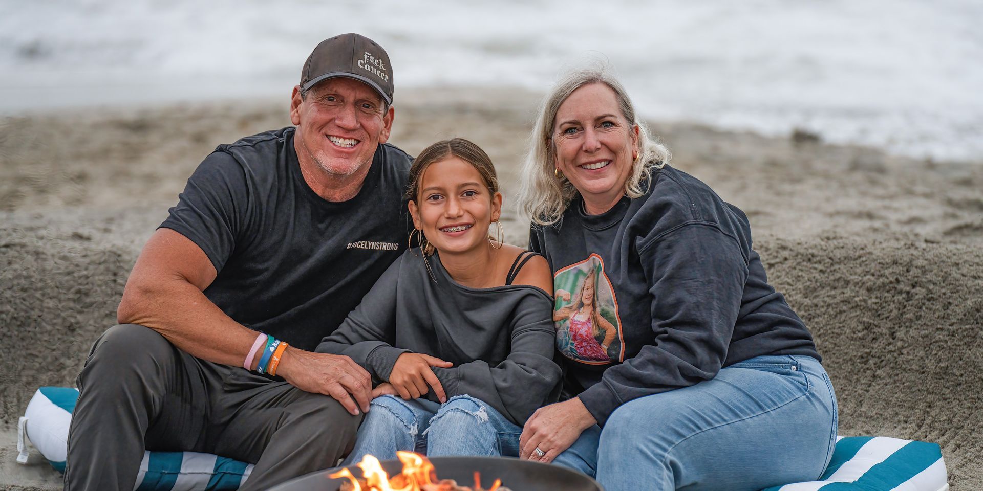 A family of three smiles while sitting near a fire pit on a beach.