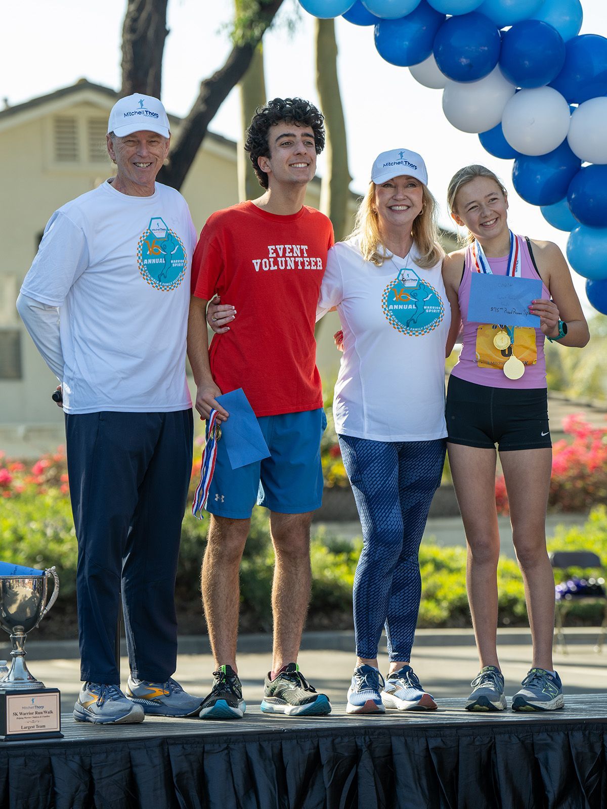 Four people on a podium with balloons. Two men, and two women. One is holding medals.
