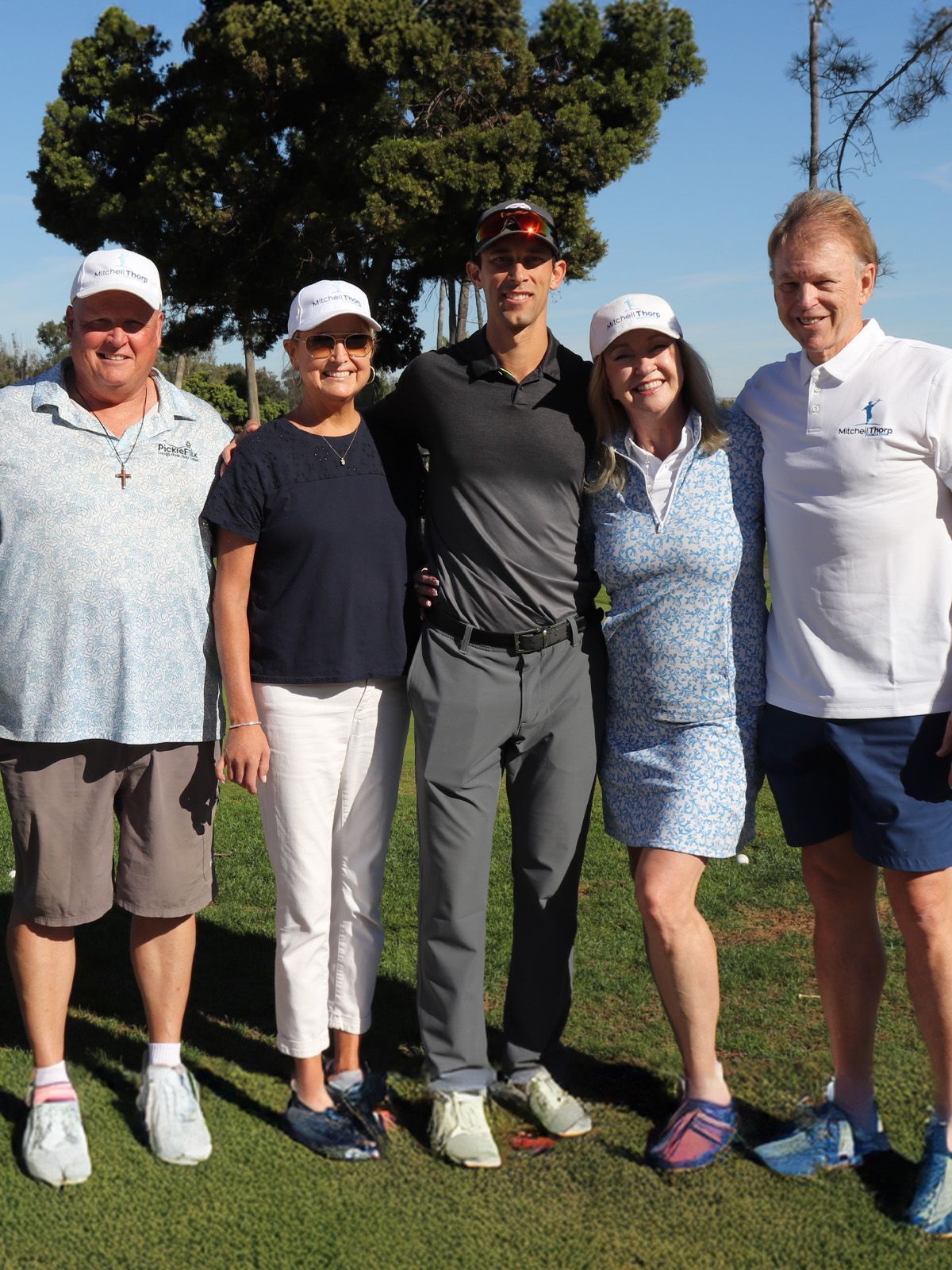 Five people smiling, standing on a sunny golf course. Two men, three women, wearing golf attire.