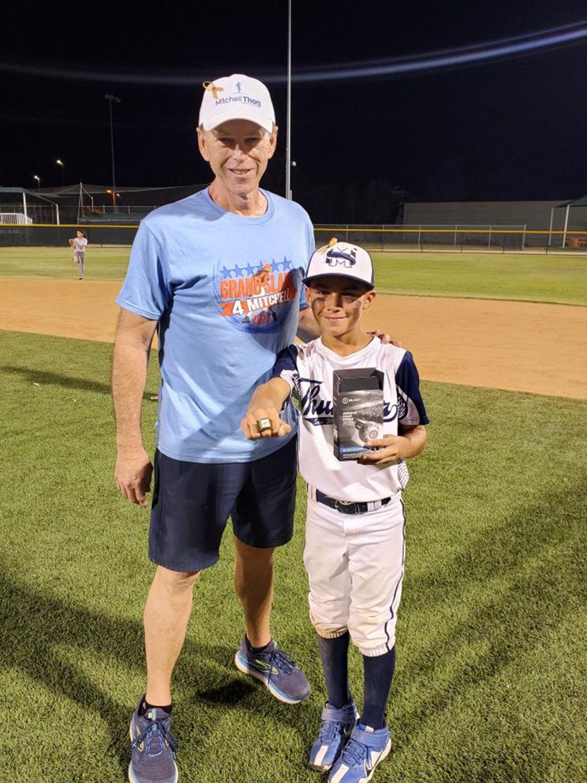 Man and young boy with trophy on a baseball field at night, both smiling.
