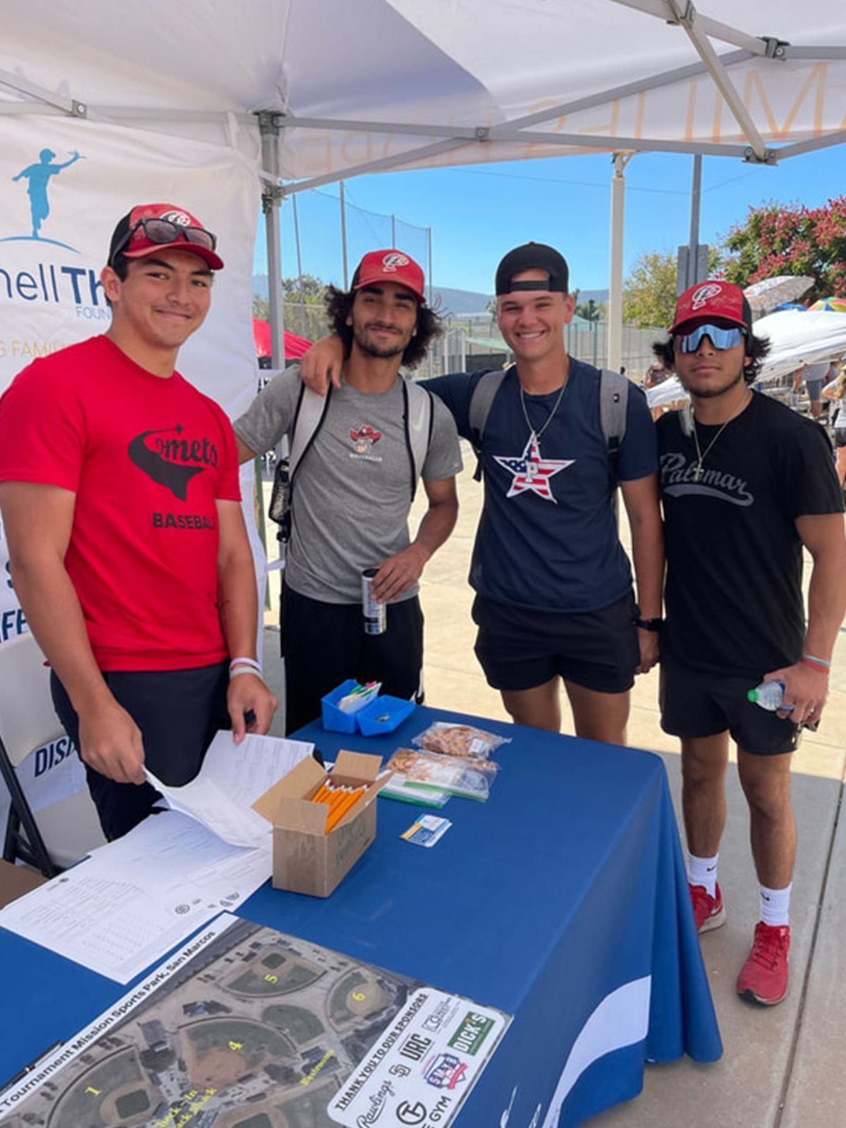 Four young men stand behind a table at an outdoor event. They wear hats, athletic clothes, and smile.