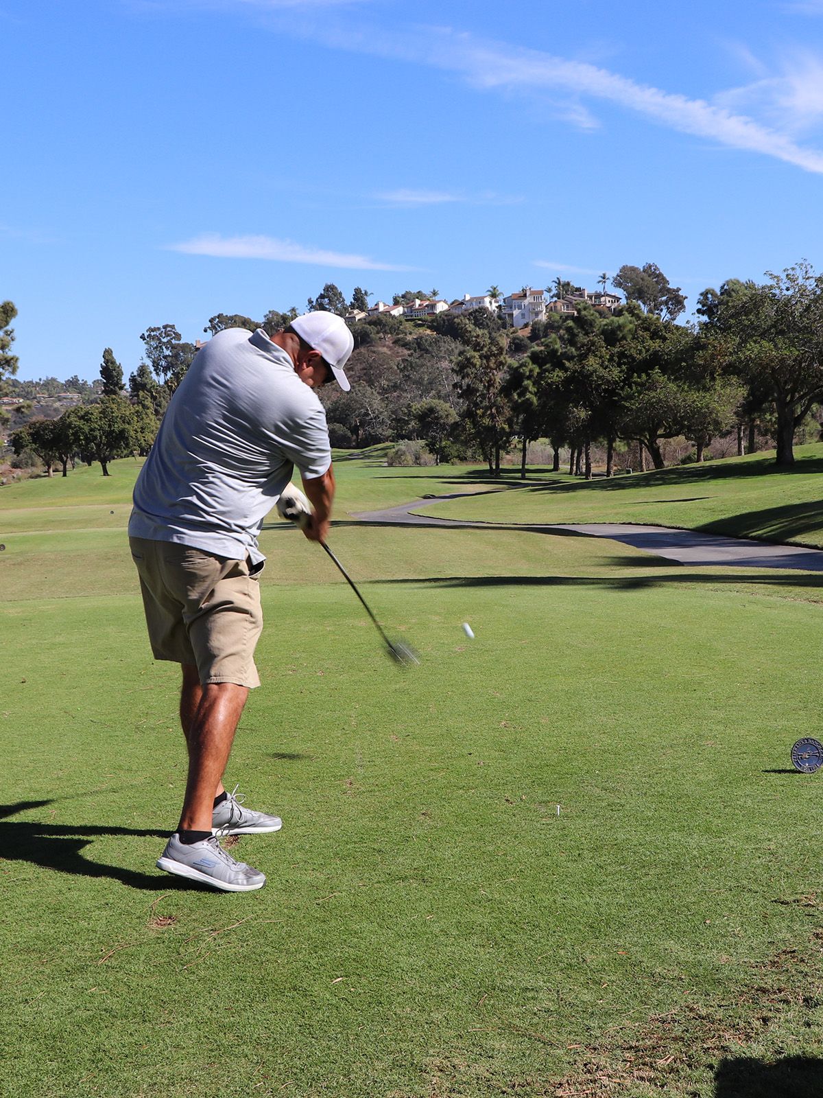 Man in gray shirt and shorts swings a golf club on a green course under a blue sky.