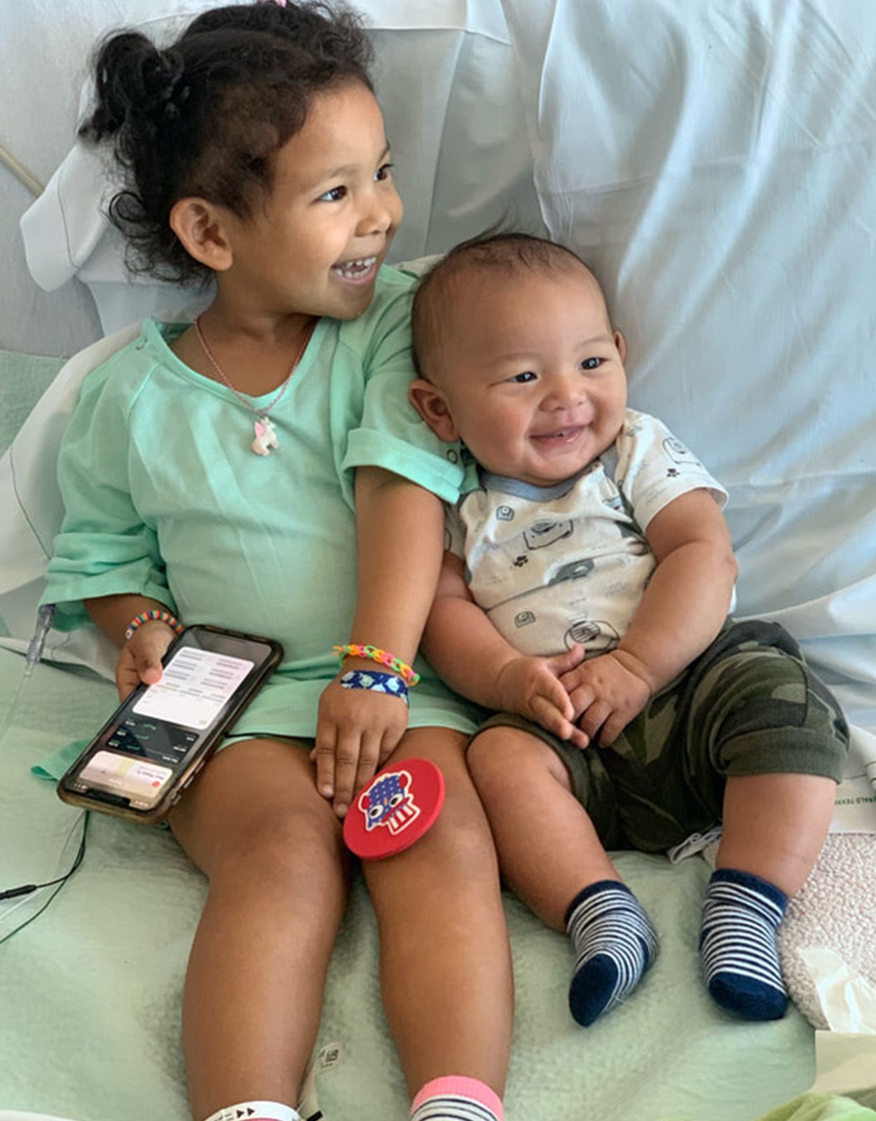 Two young children smile and embrace on a hospital bed.