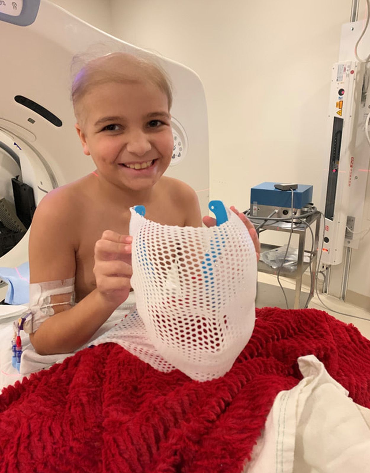 Young smiling child with head shaved, holding a radiation mask in a medical setting.