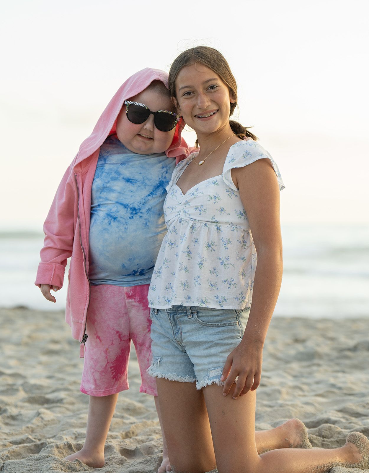 Two people smiling at the beach; one in pink hoodie and sunglasses, the other in white top and denim shorts.